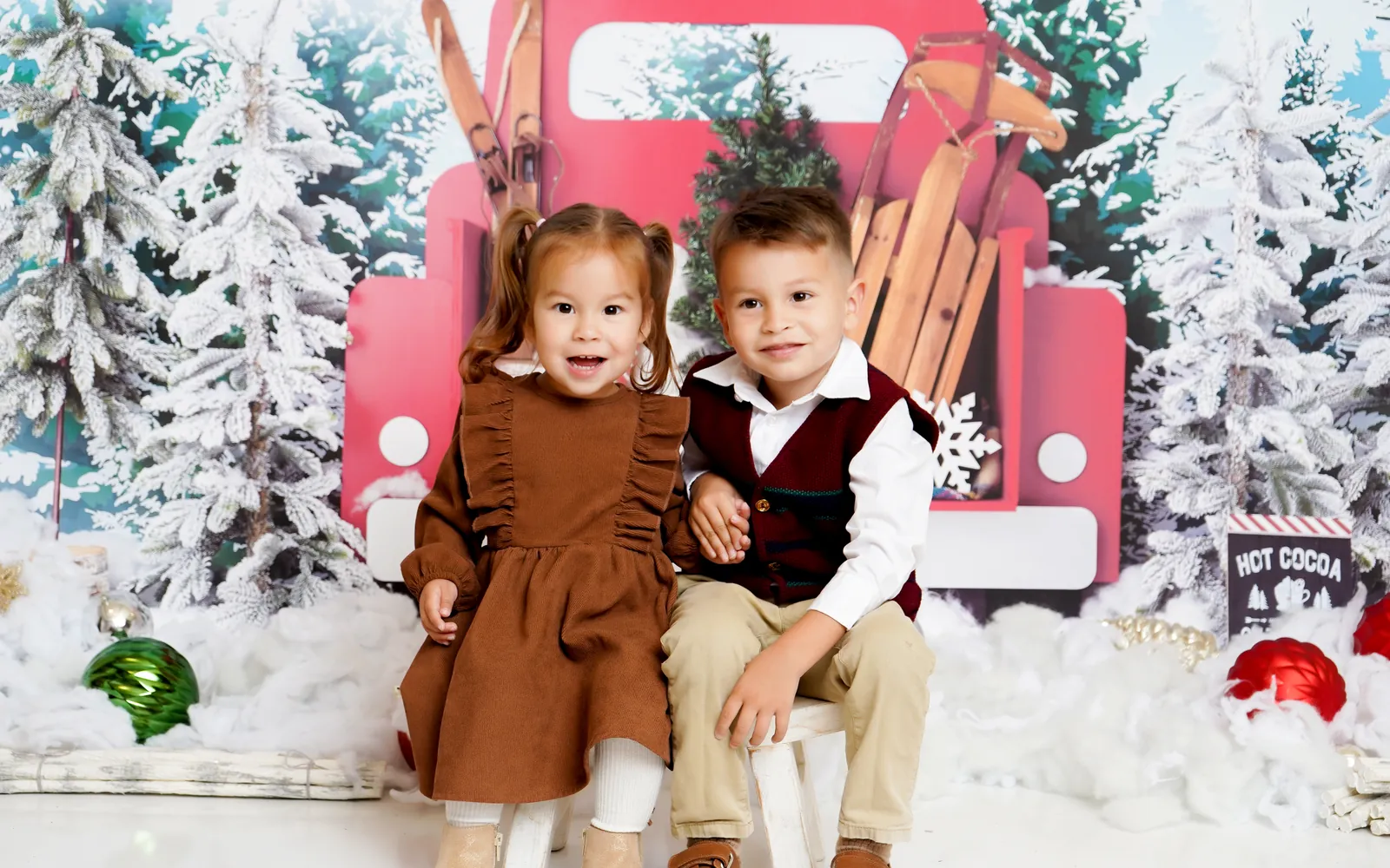 A brother and sister sitting in front of the Little Red Truck among snowy pines
