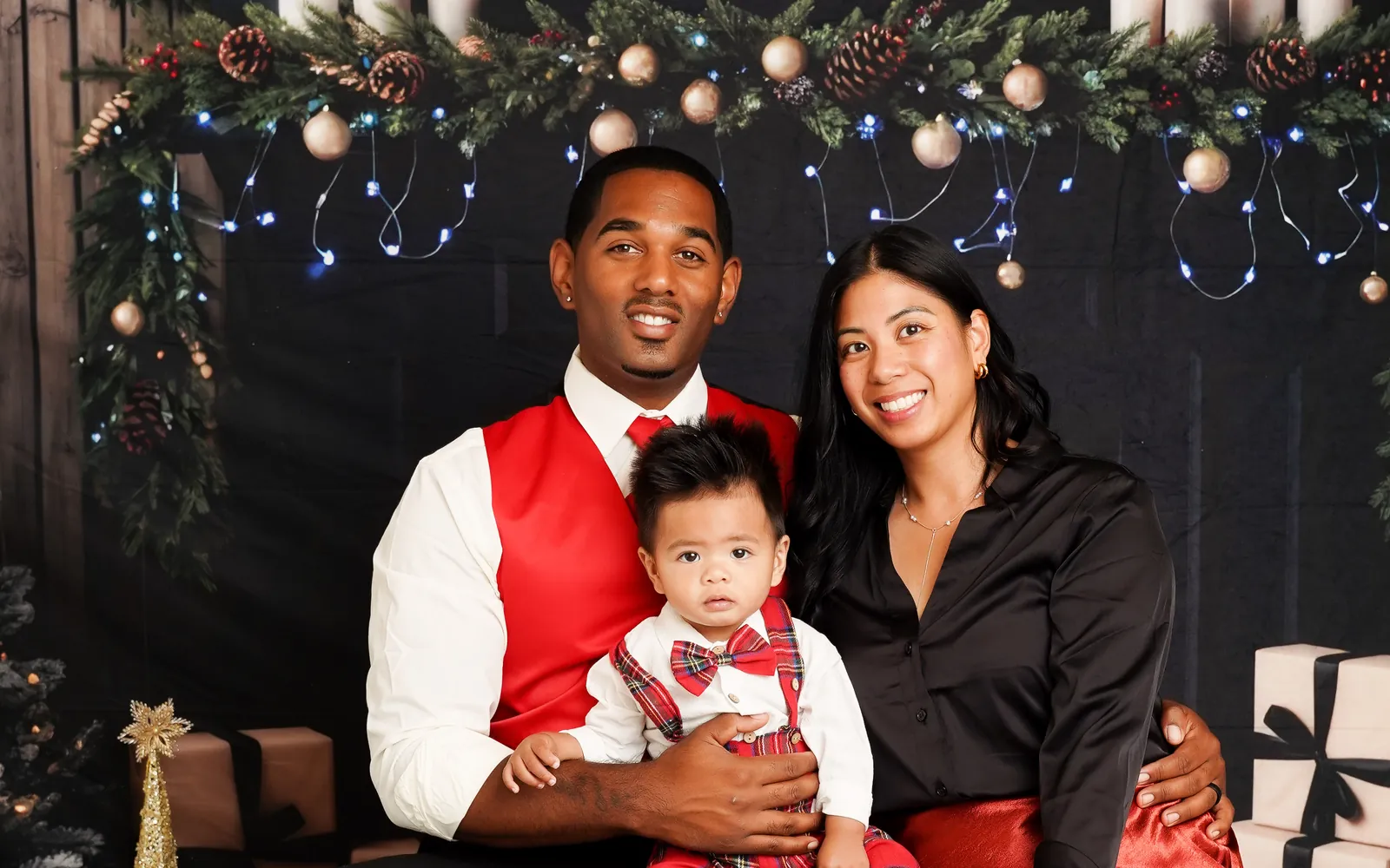 A mom and dad holding their baby beneath the candlelit garland on the Christmas Fireside set