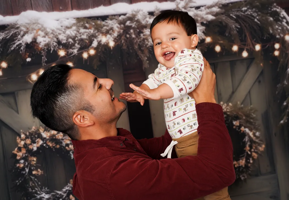 A dad lifting his laughing baby into the air on the Cozy Christmas Barn set at LaLa Holiday HQ