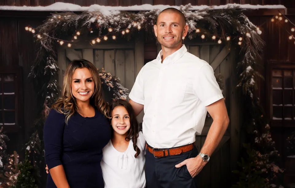 A family of three standing beneath the string lights in front of the Cozy Christmas Barn doors, dressed in wreaths and snowy garland