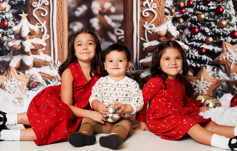 Three siblings in red sitting in the snow beneath the Gingerbread Lane archway, with frosted trees and candy ornaments all around