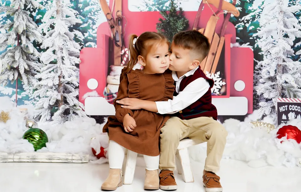 A brother giving his sister a kiss on the cheek in front of the Little Red Truck, loaded with skis and a tree among snowy pines