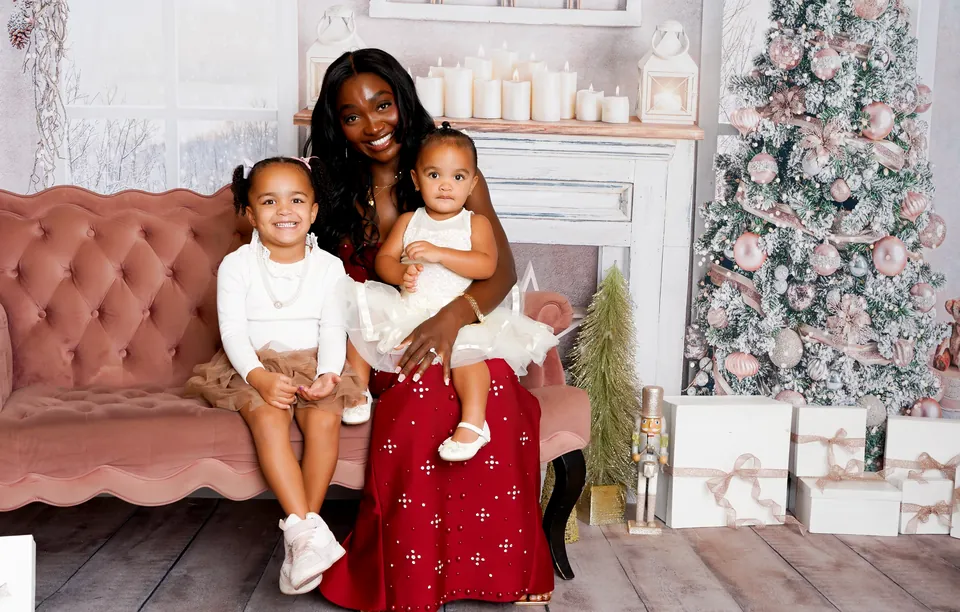 A mom with her two daughters on the pink velvet settee of the Sugarplum Christmas set, beside the candlelit mantel and blush-trimmed tree